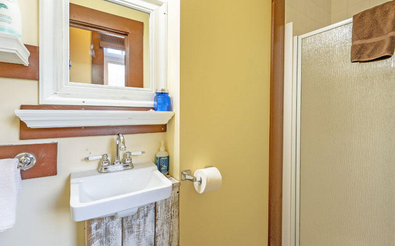 Herridge-Suite-Bathroom-at-Victoria-Falls-House-Inn-Nelson-BC Compact bathroom in the Herridge Suite at Victoria Falls House Inn, Nelson, BC, featuring a vintage-style white pedestal sink, a wood-framed mirror with a small shelf, and a walk-in shower.