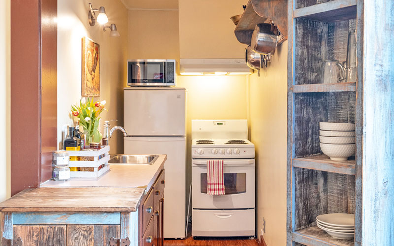 Herridge-Suite-Fully-Equipped-Kitchen-at-Victoria-Falls-House-Inn-Nelson-BC Fully equipped kitchen in the Herridge Suite at Victoria Falls House Inn, Nelson, BC, featuring a white stove, refrigerator, rustic wood shelving with dishes, and a reclaimed wood sink vanity.