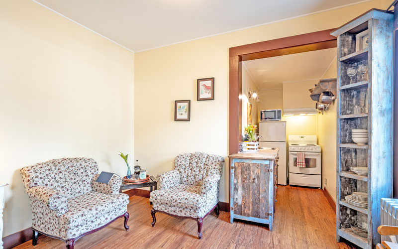 Herridge-Suite-Kitchen-Area-at-Victoria-Falls-House-Inn-Nelson-BC Cozy sitting area and kitchenette entrance in the Herridge Suite at Victoria Falls House Inn, Nelson, BC, featuring two floral armchairs, rustic wood cabinetry, and a glimpse into the fully equipped kitchen.