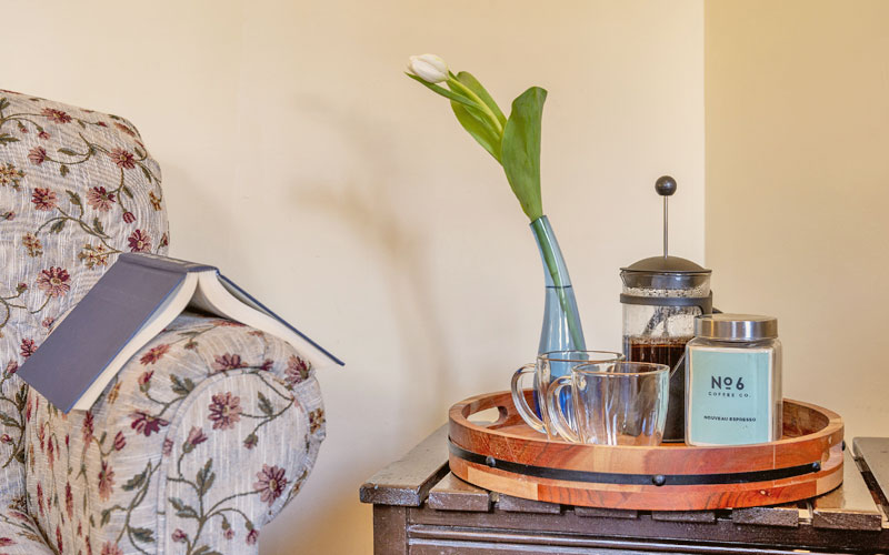 Herridge-Suite-Morning-Coffee-Victoria-Falls-House-Inn-Nelson-BC A relaxing morning coffee setup in the Herridge Suite at Victoria Falls House Inn, featuring a French press, local No. 6 Coffee Co. espresso, and a book on a side table next to a floral armchair.