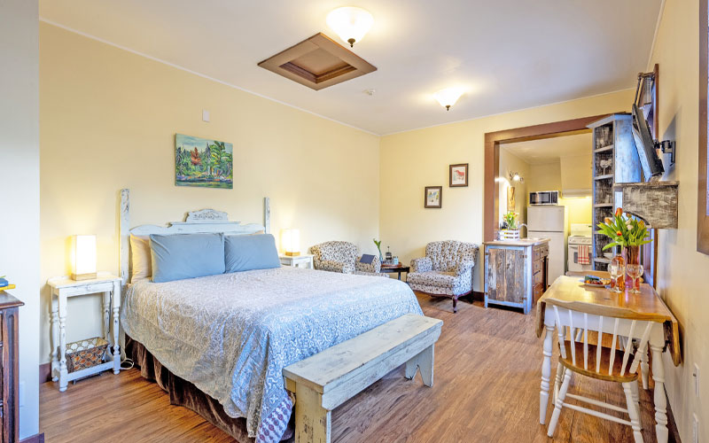 Herridge-Suite-Sleeping-Area-at-Victoria-Falls-House-Inn-Nelson-BC The open-concept studio layout of the Herridge Suite at Victoria Falls House Inn, showing a queen bed with light blue bedding, a rustic wooden bench, floral armchairs, and a dining table.